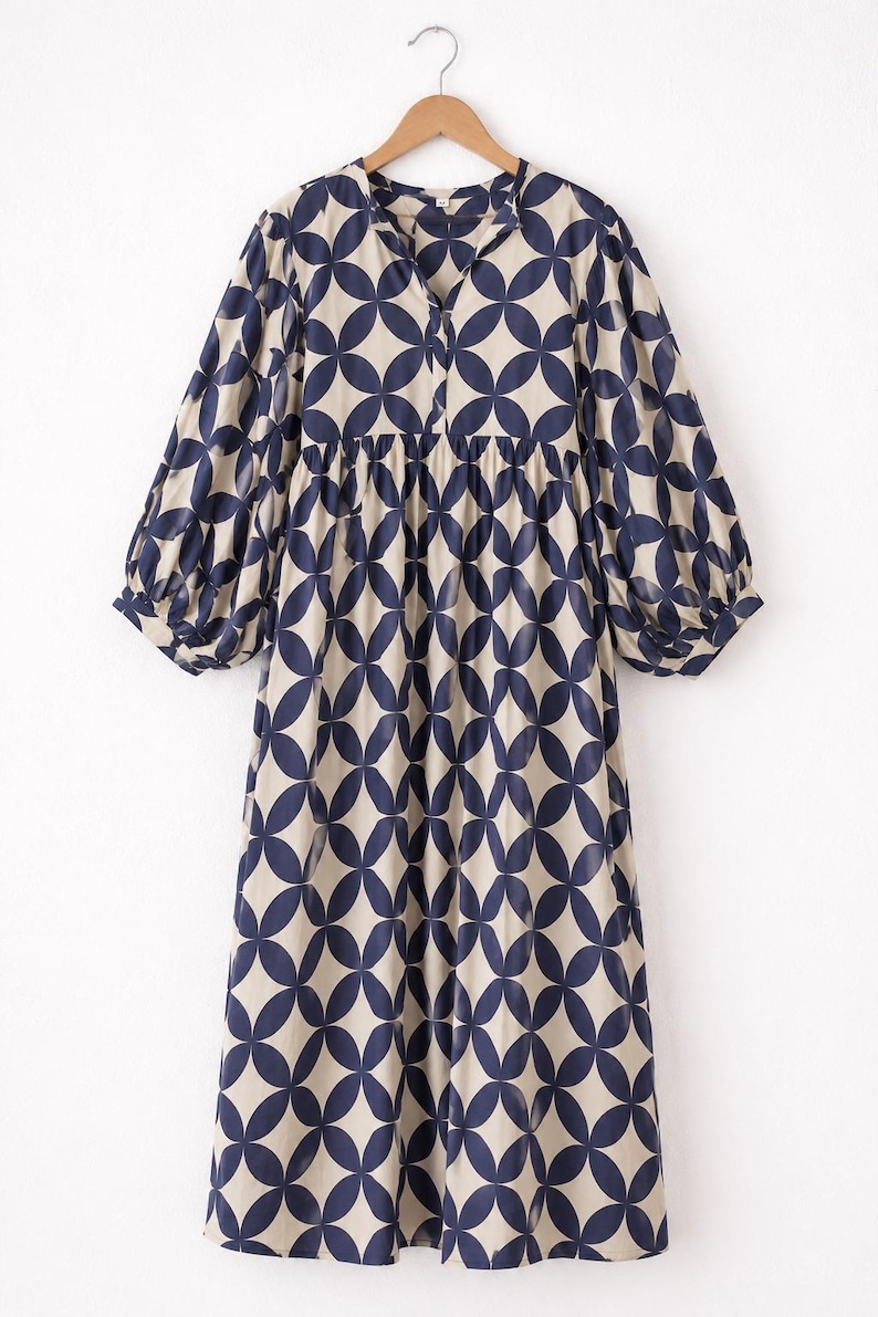 May include: A midi-length dress featuring a beige base with a repeating dark blue geometric petal pattern. The dress has a split V-neckline, a gathered waistline, and long, voluminous sleeves with elasticized cuffs, all displayed on a wooden hanger.