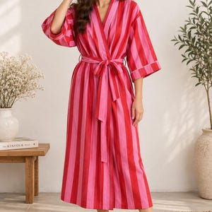 Puede incluir: Una bata hasta la pantorrilla con un llamativo patrón de rayas verticales en tonos alternos de rojo brillante y rosa vibrante. La prenda incluye mangas tres cuartos, un escote en V y un cinturón de tela a juego en la cintura.
