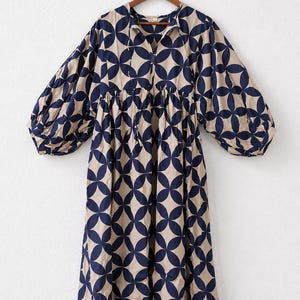 May include: A midi-length dress featuring a beige base with a repeating dark navy blue geometric circular pattern. The garment has a V-neckline with thin tie closures, a gathered waistline, and voluminous long sleeves with elasticized cuffs.