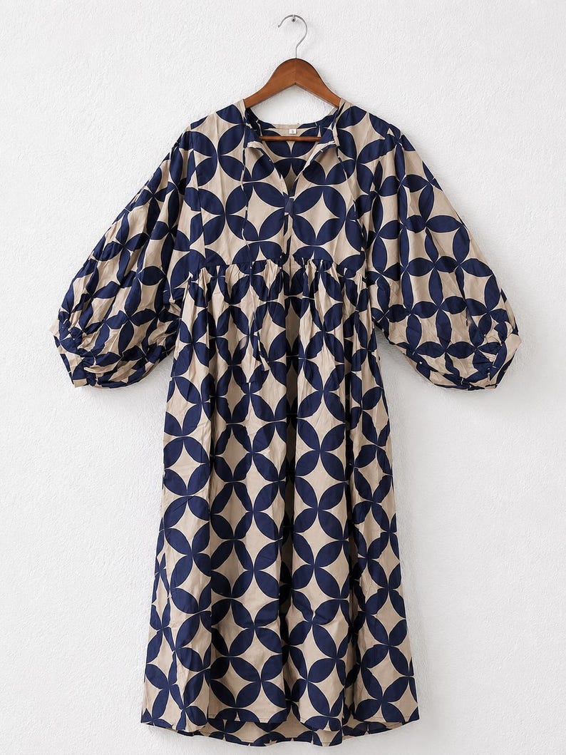 May include: A midi-length dress featuring a beige base with a repeating dark navy blue geometric circular pattern. The garment has a V-neckline with thin tie closures, a gathered waistline, and voluminous long sleeves with elasticized cuffs.