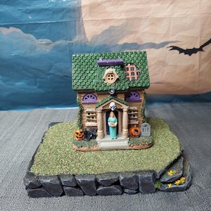 Op de afbeelding: Een miniatuur Halloween-huisje met een groen pannendak, een schedel boven de deuropening en een figuurtje in de ingang. Het huis is versierd met twee oranje pompoenen, een grafsteen met de tekst R.I.P. en ramen die met planken zijn dichtgetimmerd.