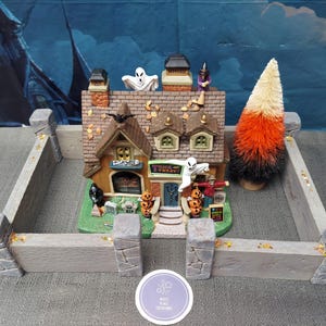 Pode incluir: Uma casa em miniatura com tema de Halloween com uma cerca de pedra. A casa tem um telhado castanho, um letreiro "Trick or Treat" e é decorada com fantasmas, abóboras e uma bruxa. Uma árvore colorida de milho doce está por perto. A cena é ambientada em um fundo azul escuro.