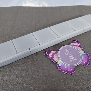 Pode incluir: Um objeto retangular cinza claro com seis seções quadradas, possivelmente um bloco de construção ou um elemento decorativo. Um adesivo de borboleta roxa com um rótulo circular que diz "Magic Place Creations" está ao lado.