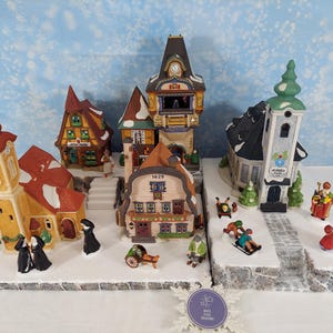 Pode incluir: Uma cena de vila de Natal em miniatura com vários edifícios com telhados vermelhos e castanhos, incluindo uma igreja e uma torre do relógio. Pequenas figuras de pessoas estão espalhadas pela cena, sobre uma base de neve.