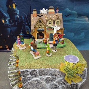 May include: A miniature Halloween village display featuring a two-story house with a Trick or Treat sign, ghosts, and pumpkins. The scene includes various figures of people and pets on a green base with a stone-textured side and a single maple leaf.