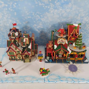 Könnte beinhalten: Zwei Weihnachtsdorfgebäude mit roten, grünen und weißen Akzenten. Eines hat eine rote Fahne, das andere einen Weihnachtsbaum. Miniaturfiguren fahren Schlittschuh und fahren Schlitten auf einer blau-weißen Basis.