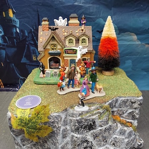 May include: A miniature Halloween village display featuring a tan house with a Trick or Treat sign, ghosts, and pumpkins. The scene includes several small figures in costumes, a candy corn colored tree, and a rocky base with a single maple leaf.