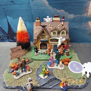 May include: A Halloween village scene featuring a house with ghosts, pumpkins, and trick-or-treaters. A welcome sign and a candy corn tree are also present. The scene includes miniature figures of people and a dragon.