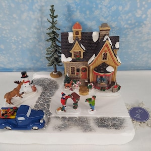 Pode incluir: Uma cena de vila de Natal com uma casa de dois andares, uma picape azul e figuras de pessoas e animais. A casa tem um telhado marrom e paredes amarelas. Um boneco de neve e uma árvore de Natal também estão presentes.