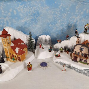 Pode incluir: Uma cena de vila de Natal em miniatura com montanhas cobertas de neve, edifícios e figuras. Os edifícios incluem uma igreja e uma casa com o número 1629. Esquiadores e outras figuras estão espalhados pela cena.