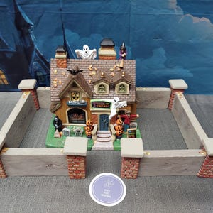 Pode incluir: Uma casa em miniatura com tema de Halloween com telhado castanho, rodeada por uma cerca cinzenta com pilares de tijolo. A casa apresenta um letreiro "Trick or Treat", fantasmas e abóboras. Um morcego está no telhado.