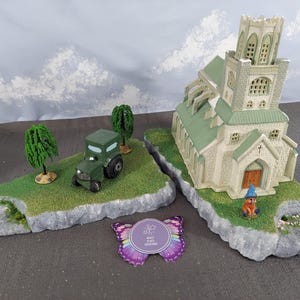 Pode incluir: Diorama em miniatura com uma igreja e um trator em plataformas rochosas separadas com relva verde e árvores. Uma etiqueta em forma de borboleta roxa está em primeiro plano. A igreja tem um telhado verde e uma torre alta.