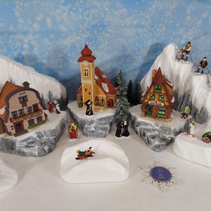 Pode incluir: Uma cena de vila de inverno com edifícios em miniatura, figuras e esquiadores em montanhas nevadas. Os edifícios incluem uma igreja, uma casa com o número 1629 e uma loja. As figuras retratam pessoas em trajes de inverno.