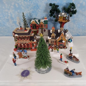 Pode incluir: Uma cena de vila de Natal em miniatura com vários edifícios, uma árvore de Natal e figuras de pessoas. Os edifícios têm exteriores vermelhos, verdes e castanhos. A árvore é verde e as figuras estão vestidas com roupas de inverno. Uma estrela está no topo da árvore.