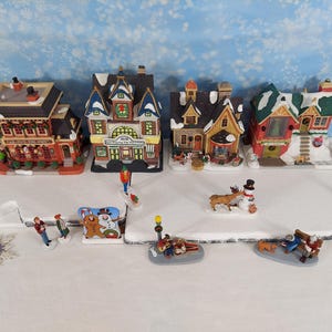 Pode incluir: Uma coleção de quatro edifícios em miniatura de uma vila de Natal e várias figuras pequenas, incluindo pessoas, um boneco de neve, uma rena e um banco, dispostos sobre uma base branca. Os edifícios apresentam telhados cobertos de neve.
