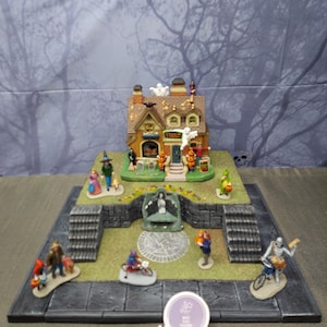 Pode incluir: Uma cena detalhada de uma vila de Halloween com uma casa de dois andares com fantasmas e abóboras. Figuras em miniatura de pessoas a pedir doces, um esqueleto de bicicleta e outros personagens estão espalhados pela casa. A cena é montada numa plataforma cinzenta.