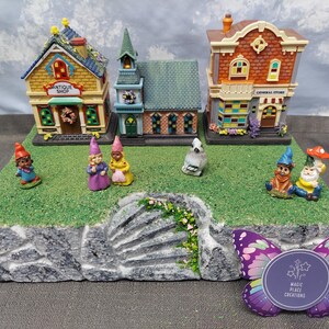 Pode incluir: Uma cena de vila em miniatura com três edifícios: uma loja de antiguidades, uma igreja e uma loja geral. Várias figuras de gnomos estão espalhadas sobre a base verde gramada. Uma etiqueta em forma de borboleta com o texto "MAGIC PLACE CREATIONS" está em primeiro plano.