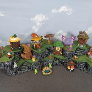 Pode incluir: Uma cena de jardim de fadas com casas em miniatura, gnomos e decorações de cogumelos. A cena inclui um lago e uma placa que diz "Bem-vindo". A cena é montada sobre uma base em camadas que se assemelha a pedra.