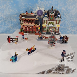 Pode incluir: Uma cena de vila de Natal em miniatura com edifícios, figuras e uma árvore decorada. Os edifícios têm fachadas detalhadas, e as figuras estão envolvidas em várias atividades de inverno. A cena é montada sobre uma base de neve com um fundo azul.