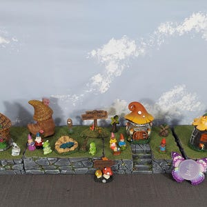 Pode incluir: Uma cena de jardim de fadas em miniatura com várias casas pequenas, gnomos e elementos decorativos. As casas têm designs de telhado de cogumelo e colmo. A cena é montada em uma base semelhante a pedra com um fundo de céu.