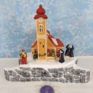 Pode incluir: Uma igreja em miniatura com telhado vermelho e paredes amarelas sobre uma base de pedra coberta de neve. Figuras de São Nicolau e três freiras com hábitos pretos estão posicionadas ao redor do edifício. Um ornamento de floco de neve branco com uma etiqueta roxa com a inscrição Magic Place Creations está colocado em primeiro plano.