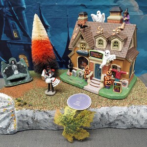 May include: A Halloween-themed miniature village scene featuring a house with a "Trick or Treat" sign, ghosts, pumpkins, and a witch. A candy corn tree and figurines of a skeleton and a couple are also present. The scene is set on a stone base.