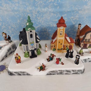Pode incluir: Um conjunto de aldeia de inverno em miniatura com uma igreja de torre verde, uma capela amarela e uma casa de telhado castanho. A cena inclui bases cobertas de neve, pequenas figuras de pessoas a andar de trenó, a caminhar e um boneco de neve.