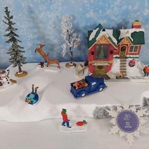 May include: A festive Christmas village display featuring a red house with a green roof, a blue truck, and miniature figures. The scene includes a snowman, deer, and people, all set on a snowy base, creating a winter wonderland.