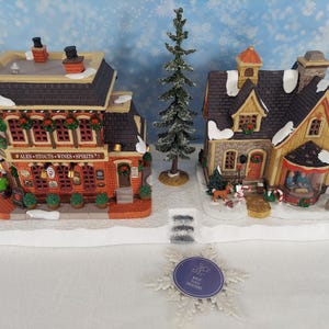 Könnte beinhalten: Eine Weihnachtsdorf-Szene mit zwei detaillierten Gebäuden, einem Pub und einem Haus, mit einem kleinen Nadelbaum dazwischen. Der Pub trägt die Aufschrift "ALES STOUTS WINES SPIRITS". Das Haus hat ein gebogenes Fenster und einen Schornstein. Die Szene befindet sich auf einer schneebedeckten Basis.