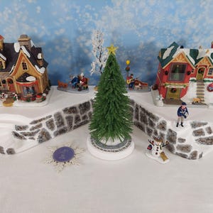 Pode incluir: Uma cena de vila de Natal com duas casas detalhadas com telhados cobertos de neve e elementos decorativos. Uma árvore de Natal verde com uma estrela dourada está no centro. Figuras em miniatura de pessoas e um boneco de neve completam a cena festiva.