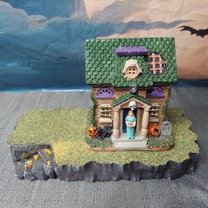 Op de afbeelding: Een miniatuur spookhuisfiguur met een groen pannendak, dichtgetimmerde ramen en een veranda met een figuur in de deuropening. De basis bevat een grasveld, twee oranje pompoenen, een zwarte kat en een grijze grafsteen.