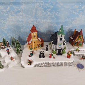 Pode incluir: Uma vila de inverno em miniatura com três edifícios pintados, incluindo uma igreja amarela, uma estrutura cinzenta com telhado verde e uma casa castanha. A cena inclui pequenas figuras de pessoas a andar de trenó, a caminhar e árvores.