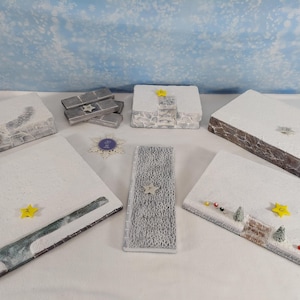 May include: A collection of rectangular and square decorative plaques featuring textured white surfaces resembling snow, accented with small yellow stars, miniature evergreen trees, and colorful beads, alongside a stack of grey rectangular blocks.