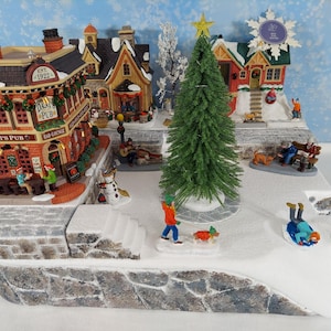 Puede incluir: Una escena de pueblo navideño en miniatura con edificios, un árbol de Navidad y figuras. La escena incluye un pub con el texto "Dani's Pub". La escena representa un paisaje invernal nevado.