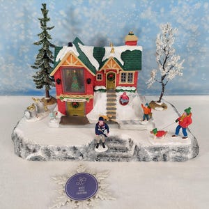 Pode incluir: Uma cena de vila de Natal em miniatura com uma casa vermelha com telhado verde e detalhes cobertos de neve. Figuras de pessoas e cães estão espalhadas ao redor da casa. A casa tem uma árvore de Natal na janela e a inscrição "Merry Christmas".