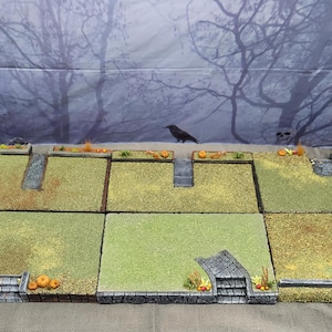 May include: A set of modular tabletop gaming terrain tiles featuring green grassy surfaces, stone-textured pathways, small staircases, and miniature orange pumpkin decorations arranged in a grid pattern on a flat surface.