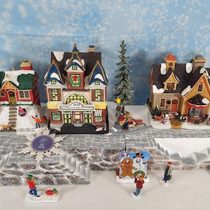 Pode incluir: Uma cena de vila de Natal com várias casas em miniatura com telhados cobertos de neve. As casas são de várias cores, incluindo vermelho, verde e castanho, e são decoradas com detalhes festivos. Pequenas figuras de pessoas e animais estão espalhadas pela cena.