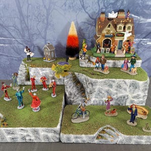 Puede incluir: Una escena de pueblo de Halloween con figuras en miniatura sobre plataformas escalonadas. La escena incluye una casa con un letrero de "Trick or Treat", figuras disfrazadas, una iglesia y un árbol de maíz dulce.