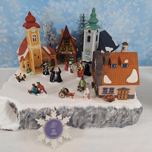 Pode incluir: Uma cena de vila de Natal em miniatura com edifícios cobertos de neve, incluindo uma igreja e casas. Pequenas figuras de pessoas estão espalhadas pela cena, algumas andando de trenó e outras caminhando. Os edifícios têm telhados vermelhos e laranja.