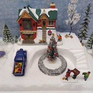 May include: A Christmas village scene featuring a red brick house with a green roof, snow-covered landscape, and miniature figures. A blue truck carries gifts, and a decorated Christmas tree stands in the center.