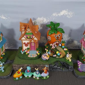 May include: A collection of Easter-themed miniature houses and figurines. The houses feature colorful designs with blue, orange, and pink roofs. Bunny and other animal figurines are placed around the houses. The houses are set on gray stone bases.