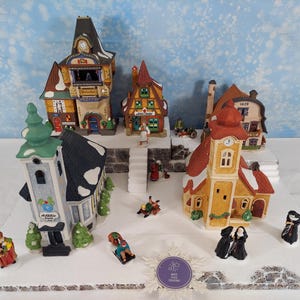Pode incluir: Uma cena de vila de Natal em miniatura com vários edifícios, incluindo uma igreja, lojas e casas. Os edifícios são pintados em várias cores com telhados cobertos de neve. Pequenas figuras de pessoas estão espalhadas pela vila.