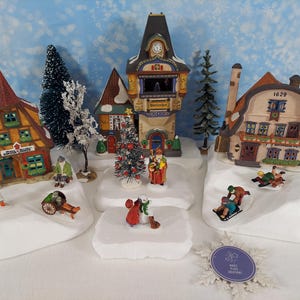 Pode incluir: Uma cena festiva de uma vila de Natal com casas em miniatura, árvores e figuras. Os edifícios têm detalhes arquitetónicos e são decorados com neve. Várias figuras estão envolvidas em atividades de inverno.