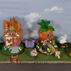 Pode incluir: Uma cena de vila de Páscoa caprichosa com casas em miniatura, uma estrutura em forma de cenoura e várias figuras de coelhos. As casas têm telhados coloridos e são cercadas por vegetação e flores. A cena é montada em uma base verde.