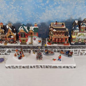 Pode incluir: Uma cena de vila de inverno com casas em miniatura, figuras e uma árvore de Natal. As casas têm telhados cobertos de neve e são decoradas com cores festivas. As figuras retratam pessoas desfrutando de atividades de inverno.