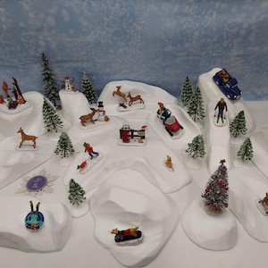 Op de afbeelding: Een winterdorpsscène met miniatuurbeeldjes van skiërs, rodelaars en een auto op besneeuwde heuvels. De scène bevat kleine kerstbomen, herten en een sneeuwpop. De achtergrond is lichtblauw met een sneeuweffect.