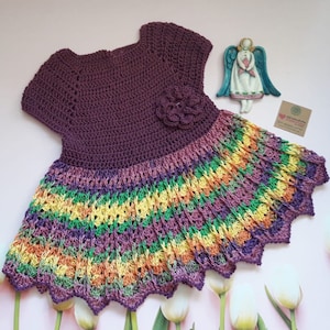 May include: A crocheted baby dress with a purple bodice and a multi-colored skirt. The skirt has stripes of yellow, green, orange, and purple.