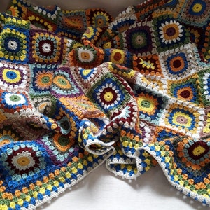 May include: A colorful crocheted blanket with a variety of squares in shades of blue, green, yellow, orange, red, and brown. The blanket is made with a granny square pattern and has a white border.