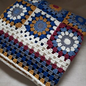 May include: A crocheted blanket with a patchwork design featuring squares in shades of blue, gray, white, burgundy, and gold.