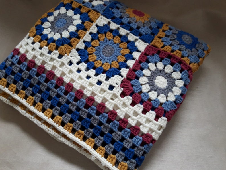 May include: A crocheted blanket with a patchwork design featuring squares in shades of blue, gray, white, burgundy, and gold.